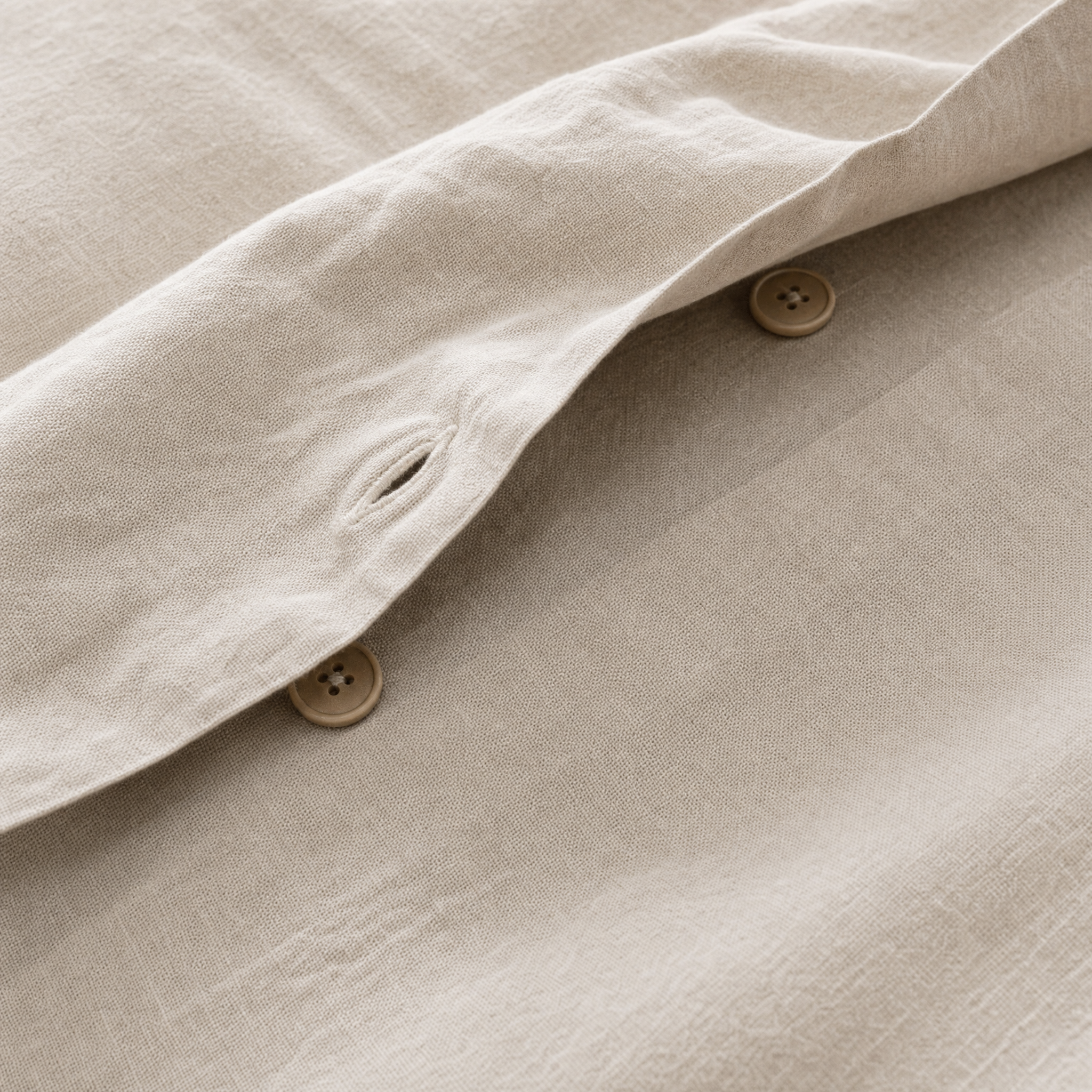 Close-up of a light beige cotton fabric coverlet with visible buttons and a folded cuff showing the texture and buttonhole detail.